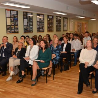 Na zdjęciu widzimy grupę osób siedzących w sali, na spotkaniu, seminarium lub prezentacji. Pokój ma drewnianą podłogę i ściany ozdobione oprawionymi fotografiami i certyfikatami. Uczestnicy są ubrani w formalne i półformalne stroje, a niektórzy trzymają notatniki lub papiery, co sugeruje, że jest to profesjonalne lub edukacyjne spotkanie. Oświetlenie jest jasne, a sala wydaje się dobrze zorganizowana z rzędami krzeseł dla publiczności.