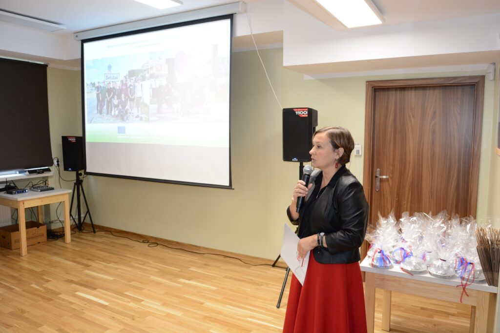 Na zdjęciu znajduje się osoba przemawiająca do mikrofonu w sali konferencyjnej. W tle widoczny jest ekran projekcyjny z wyświetlonym slajdem, na którym znajdują się zdjęcia grupowe oraz logotypy programów Erasmus+ i Europejskiego Funduszu Społecznego. Po lewej stronie ekranu znajduje się stół z laptopem i głośnikiem, a po prawej stronie stołu widoczne są zapakowane prezenty lub upominki na stole. W tle widać również zamknięte drzwi.