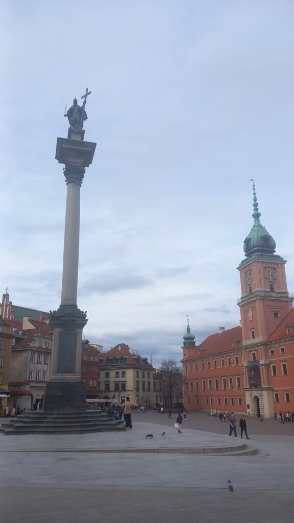 Zdjęcie przedstawia Plac Zamkowy w Warszawie. Na pierwszym planie widoczna jest Kolumna Zygmunta III Wazy, jedna z najbardziej rozpoznawalnych warszawskich budowli. W tle znajduje się Zamek Królewski,