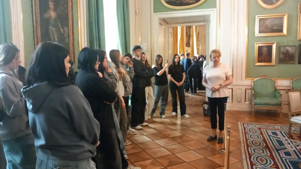 Zdjęcie przedstawia grupę młodzieży zwiedzającą Zamek Królewski w Warszawie. Uczestnicy słuchają przewodniczki w eleganckim, zabytkowym wnętrzu z zielonymi ścianami, obrazami w złotych ramach