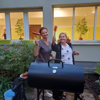 Na zdjeciu widać dwie uśmiechniete wychowawczynie przy grillu w ogrodzie bursy. Trzymają kiełbaski i szczypce, obok na talerzu leży więcej jedzenia. W tle widać duże okna z roślinami na parapecie. Atmosfera jest luźna i towarzyska.