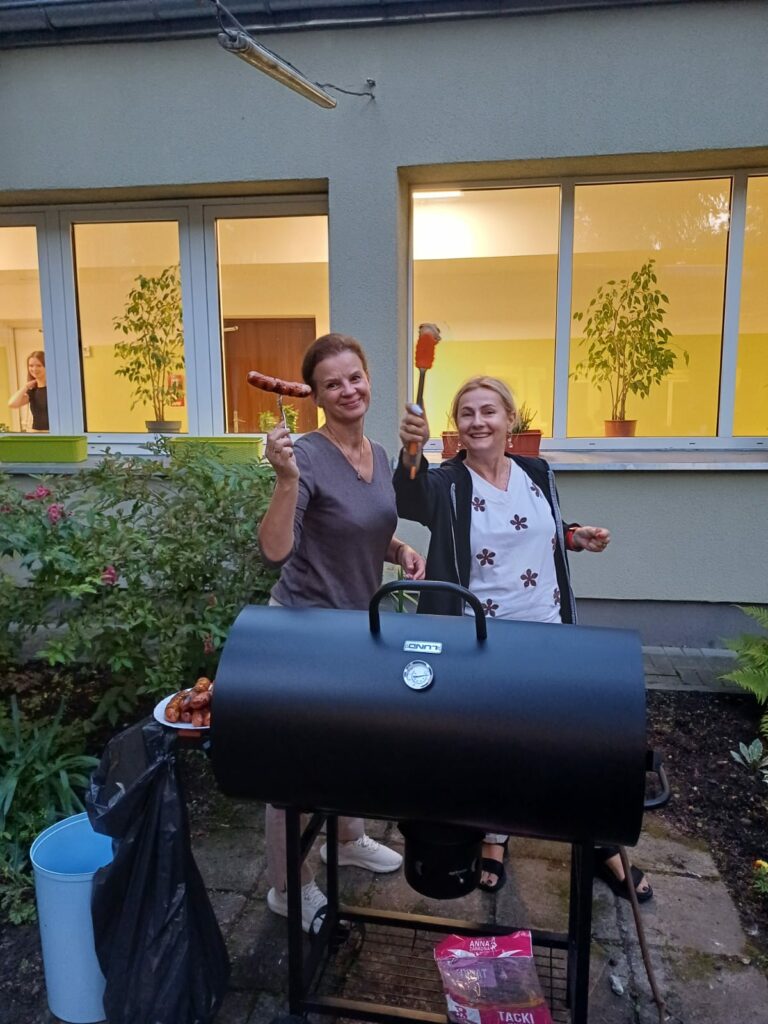 Na zdjeciu widać dwie uśmiechniete wychowawczynie przy grillu w ogrodzie bursy. Trzymają kiełbaski i szczypce, obok na talerzu leży więcej jedzenia. W tle widać duże okna z roślinami na parapecie. Atmosfera jest luźna i towarzyska.