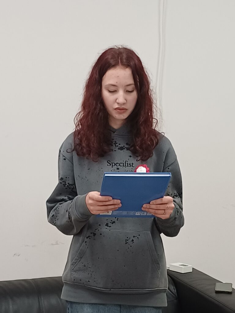 1 – Na zdjęciu widoczna jest młoda osoba stojąca w pomieszczeniu o jasnym tle, trzyma w rękach niebieską teczkę i wygląda na skupioną, jakby czytała tekst. Ubrana jest w szarą bluzę z nadrukiem oraz jasne spodnie. Za nią znajduje się ciemna kanapa i niewielki stolik.
