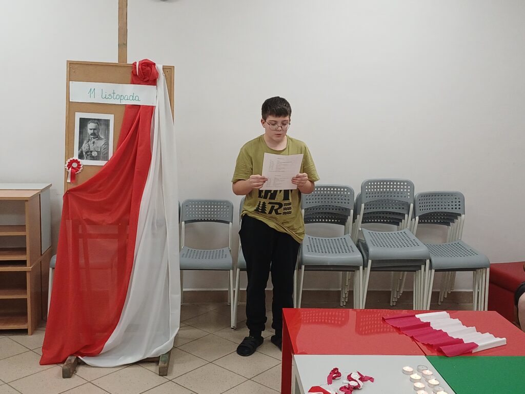 1 – Na zdjęciu widoczna jest młoda osoba stojąca w pomieszczeniu o jasnym tle, trzyma w rękach niebieską teczkę i wygląda na skupioną, jakby czytała tekst. Ubrana jest w szarą bluzę z nadrukiem oraz jasne spodnie. Za nią znajduje się ciemna kanapa i niewielki stolik.
