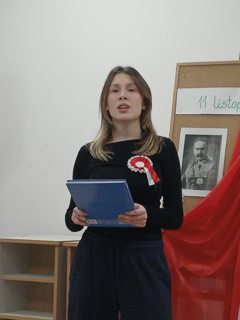 3 – Na zdjęciu widać młodą osobę stojącą przy dekoracji z okazji Święta Niepodległości. Trzyma w rękach niebieską teczkę i  wykonuje utwór muzyczny  podczas spotkania patriotycznego. Na tablicy obok znajduje się napis „11 Listopada” oraz portret postaci historycznej , a całość ozdobiona jest biało – czerwoną tkaniną.
