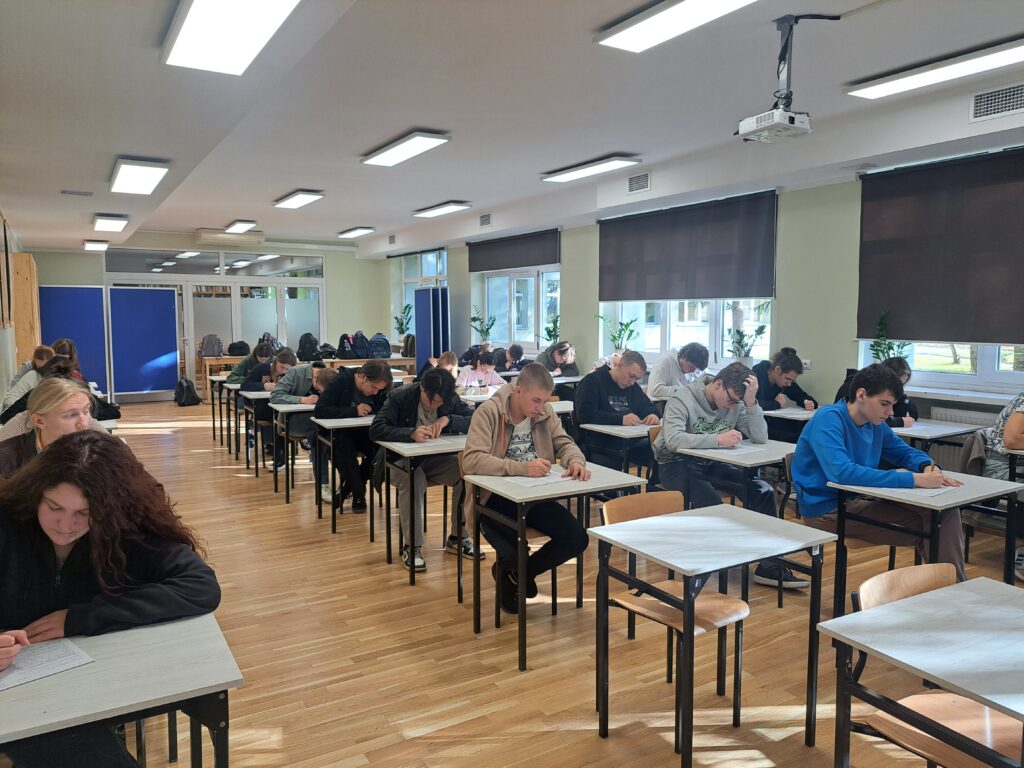 Grupa uczniów siedzi w szkolnej sali przy pojedynczych ławkach i pisze test. W pomieszczeniu jest jasno, widać okna z roletami oraz tablice po lewej stronie