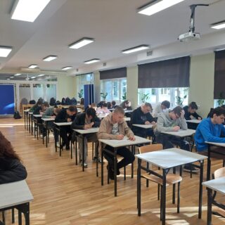 Grupa uczniów siedzi w szkolnej sali przy pojedynczych ławkach i pisze test. W pomieszczeniu jest jasno, widać okna z roletami oraz tablice po lewej stronie