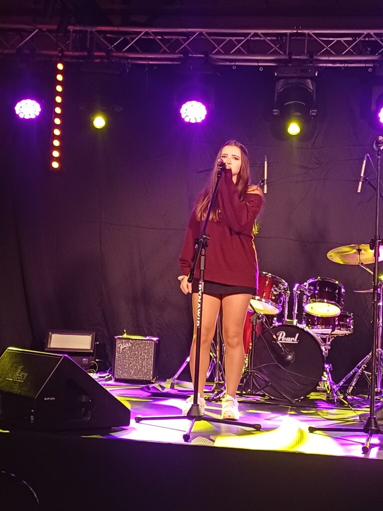 1 – Młoda wokalistka z bursy, otulona światłem sceny, porusza publiczność swoim głosem podczas festiwalu piosenki.