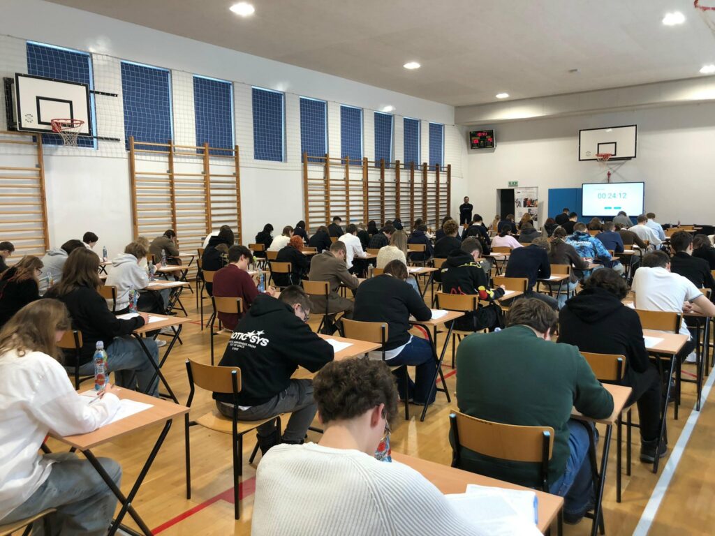 „Uczniowie siedzą w sali gimnastycznej przy pojedynczych stolikach i w skupieniu piszą test konkursowy z języka angielskiego. W tle znajduje się ekran z odliczaniem czasu.”